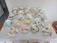 Top view of multiple teacups and saucers, all with various floral and gold-trimmed designs, arranged on a white tablecloth.