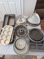 Photo shows multiple metal baking pans and trays arranged on a white tablecloth, including loaf pans, bundt cake pans, heart-shaped pans, muffin tins, round cake pans, pizza pan, and square compartment tray.