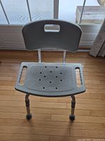 Front view of adjustable bath/shower chair showing textured seat with drain holes, backrest with handle, and rubber feet.