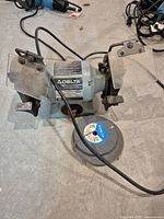 Full view of the Delta bench grinder showing two grinding wheels, safety shields, and spare grinding wheel on the floor.