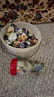 Wide shot of a plastic bucket filled with assorted buttons and a clear jar with a red lid containing various sewing items placed beside it on a carpet.