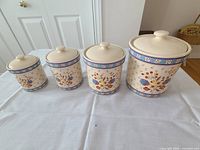 Four ceramic canisters of different sizes arranged from small to large on a white table, featuring colorful floral patterns and blue decorative bands near the top, each with a knobbed lid.