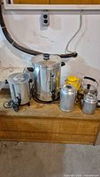 Two electric coffee percolators, large stove-top percolator, three different kettles/carafes, all metal, arranged on wooden surface.