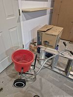Photo showing a red push-style fertilizer spreader with two black wheels and a metal frame. Nearby are a ladder, a Lowe's Medium box, gardening gloves, and other small items.