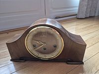 Front view of wooden mantel clock showing curved top design, round clock face with gold-tone Arabic numerals and hands behind a glass cover with gold-tone bezel.