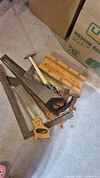 Photo shows a wooden mitre box, two hand saws with wooden handles, a hand axe, and a hammer laid on a concrete floor next to a cardboard box.