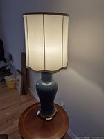 Tall green ceramic lamp with a white pleated fabric shade with gold trim on a gold-accented base, shown lit on a round wooden table.