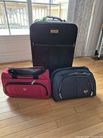 Photo showing one large black suitcase and two carry-on bags (red and dark blue) on wooden floor near windows.