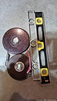 Photo shows two round metal measuring tapes, one larger and one smaller, and two spirit levels, one metal and one plastic black with yellow vials, laid flat on a concrete surface.