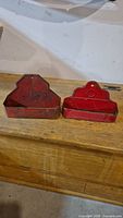 Two red antique metal holders on a wooden surface, showing both front and side views with wall mount holes and open tops.