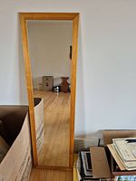 Full length mirror shown upright against a wall, reflecting a room with wooden floor and boxes.