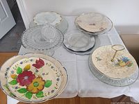 Photo showing a variety of glass and ceramic platters and serving dishes on a table including clear glass platters, scalloped edges, floral ceramic platters, and a two-tiered serving dish with a gold handle.