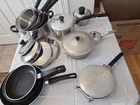Photo showing an assortment of pots, pans, lids, and a taco maker on a white surface