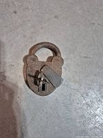 Top-down close-up view of the rusty vintage railway padlock with key inserted and visible on a rough concrete floor.