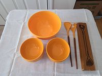 Photo showing one large orange salad bowl, four smaller matching orange bowls stacked, and a pair of wooden-handled orange plastic salad tongs placed next to them on a white tablecloth.