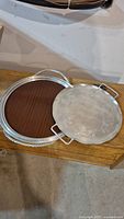 Photo showing two metal serving trays, one round engraved tray with handles on top of a larger rope edge serving tray with a wooden base insert.