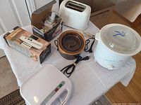 Photo showing all small kitchen appliances on a white table including two crock pots, toaster, sandwich maker, and digital food thermometer
