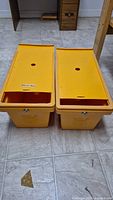 Two yellow plastic small storage boxes with sliding lids and circular holes in the lids. Positioned on tiled floor.