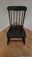 Front view of the small black wooden rocking chair showing the curved top backrest with vertical spindles and solid seat.