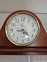 Close-up photo of clock face showing Ego brand, black Arabic numerals, black minute markers, decorative black hands and gold-tone bezel.