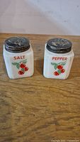 Front view of the pair showing cherry motif and red SALT and PEPPER lettering on milk glass shakers with worn metal lids.