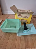 Vintage Spartan vaporizer unit with turquoise base and cover, black vaporizing element, and white power cord, beside original yellow and white box.