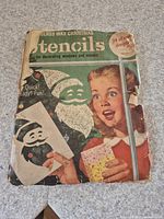 Front cover of vintage Glass Wax Christmas stencil booklet showing a child and Santa figure using stencils. Shows wear and tear on edges and corners.