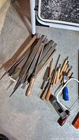 Photo showing an assortment of woodworking files, chisels, a small hand saw, scrapers, and paint scrapers on a floor surface.