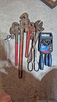 Top-down view displaying all nine tools laid on a concrete floor: three red pipe wrenches, one silver adjustable wrench, large metal scissors, and a new three-piece screwdriver set in blue packaging.