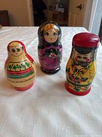 Front view of three vintage Matroshka dolls including female in yellow dress, female in black headscarf and purple dress, and male figure in yellow traditional dress with red hat on white surface.