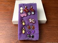 Top view of all ten pairs of semi-precious stone earrings mounted on purple foam with white box background. Shows variety of colors and styles including moonstone, amber, amethyst, and onyx.