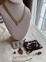 Wide gold-tone chain necklace with square clasp alongside various other jewelry pieces including earrings, pendants, and rings arranged on table.
