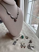 Full view of all 7 sterling silver jewelry pieces including necklace on display bust and earrings and rings laid flat with visible red and green stones.
