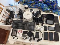 Wide view of assortment electronics including keyboard, smartphones, headphones, cables, vintage iPod, baby monitor, and other devices on a table and laundry basket.