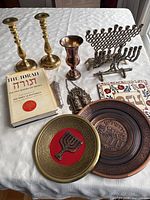 Photo showing full lot layout on white tablecloth including brass candlesticks, large brass menorah, copper Kiddush cup, Torah book, decorative plates, and mezuzah case with chain.
