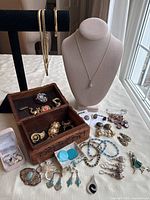 Photo of wooden treasure box filled with various vintage brooches, pins, and earrings placed next to assorted jewelry pieces including necklaces, bracelets, and earrings laid out on table.