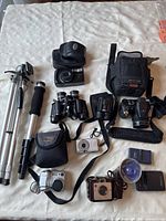 Photo showing various cameras, binoculars, tripods, monopod, and accessories on fabric surface.
