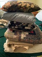 Stack of bedding items including floral pillow, light blue pillow, patchwork quilt, woven blanket, and other layered blankets.