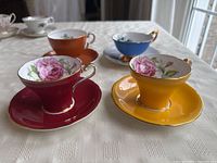 Four fine china tea cups on matching saucers in red, yellow, blue, and orange with floral interior patterns and gold trim on a white patterned tablecloth.