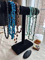 Multiple polished stone necklaces and pendants on a black jewelry stand, showing colors including black, turquoise, light blue, and brown.