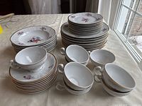 Full set of Bernadotte Sonata fine china dishes comprising stacks of bowls, cups, saucers, dessert plates, and dinner plates arranged on a table.