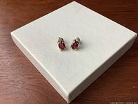 Pair of small vintage earrings featuring deep red oval garnets and round imitation diamonds set in polished 14K yellow gold on a white textured display box surface.