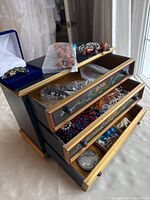 Overall view of vintage wooden jewelry box with floral detail drawers open showing various costume jewelry pieces inside and on top including beaded necklaces and bracelets in plastic bags and loose.