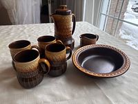 Full set showing teapot, four mugs, creamer, and serving bowl on a white tablecloth with window background. Highlights brown gradient glaze and pattern.