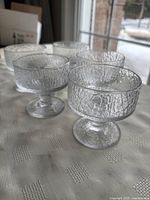 Five clear vintage Iittala dessert cups on a white tablecloth, showcasing the textured glass water droplet pattern and pedestal bases, photographed from an angle.