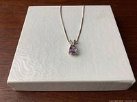 Close-up of the amethyst pendant against a textured white box background highlighting the stone's teardrop shape and silver setting.