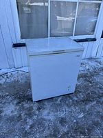 Front and left side view of the white Arctic King chest freezer placed outside on icy ground, shows wear and scuffs.