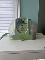 Front view of Bissell Little Green Machine in green with clear water tanks. Shows handle, control dial and hose connection.