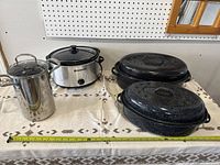 Full lot overview on table showing all items: two black speckled roasting pans, stainless steel stock pot, Bravetti Crock Pot slow cooker.