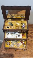 Opened wooden jewelry box displaying three tiers with assorted costume jewelry including necklaces, earrings, brooches, and other items